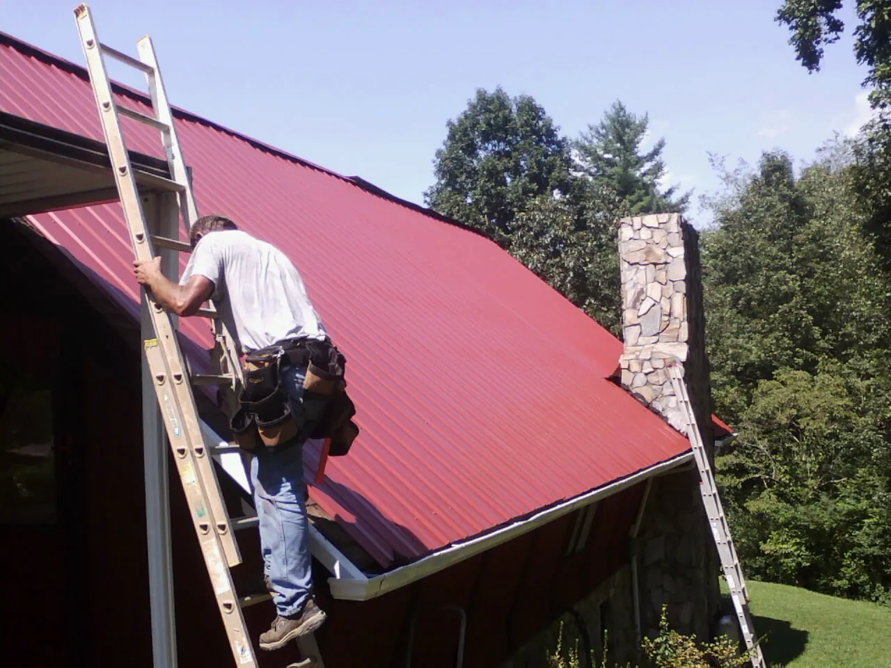 Roofer on a ladder beside a red metal roof and stone chimney — Soffit & Fascia Repair by Roofing Brothers in Hallsville